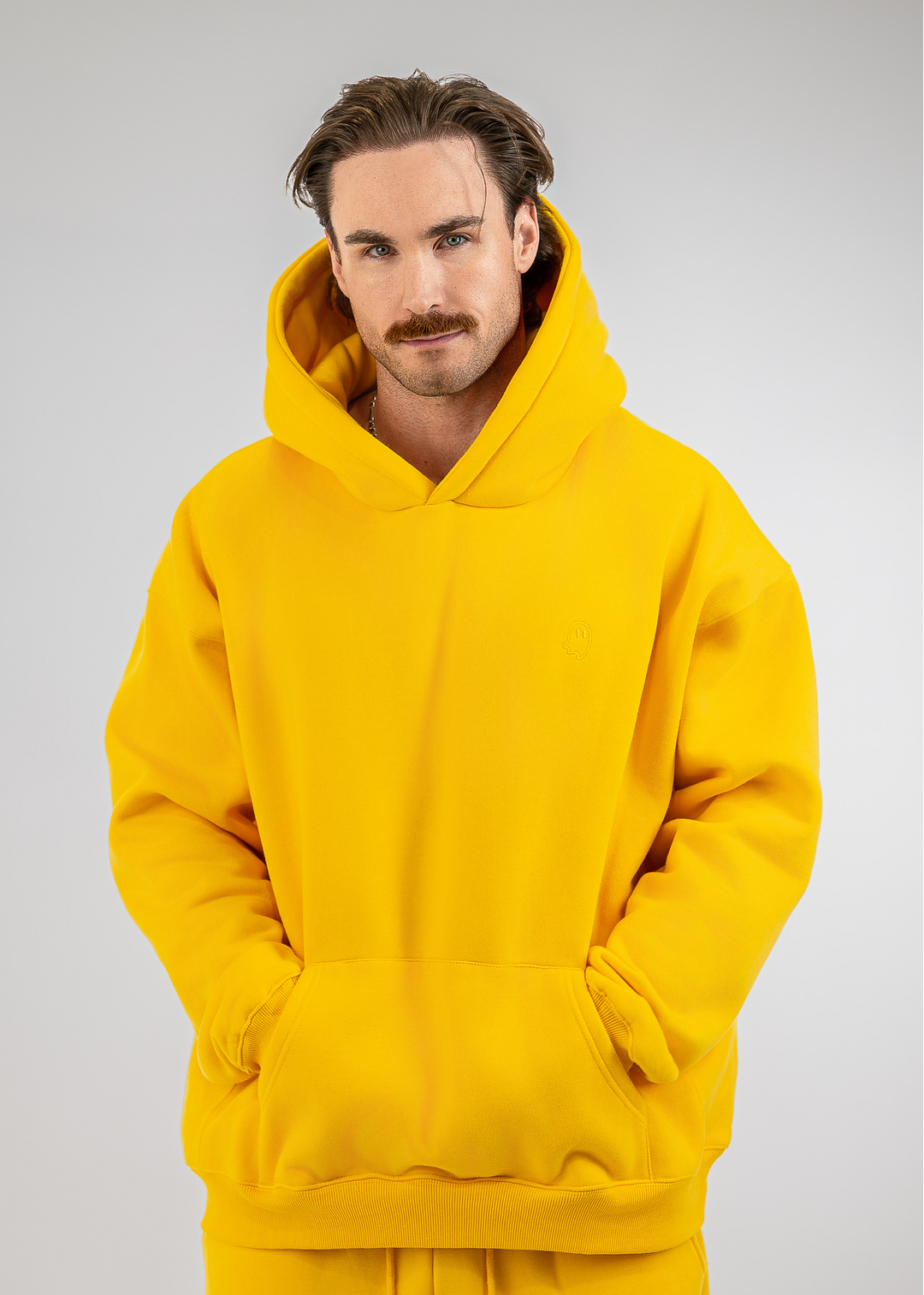 A man with a mustache wearing a bright yellow oversized pullover hoodie with a front kangaroo pocket, photographed against a neutral light gray studio background.