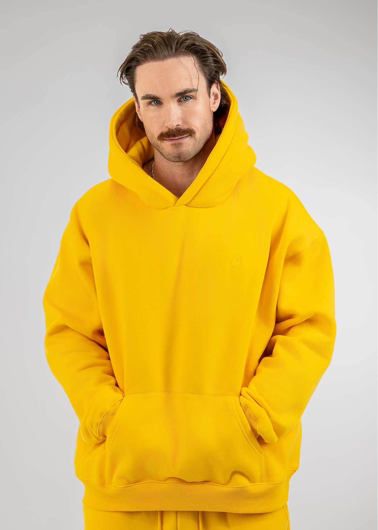 A man with a mustache wearing a bright yellow oversized pullover hoodie with a front kangaroo pocket, photographed against a neutral light gray studio background.