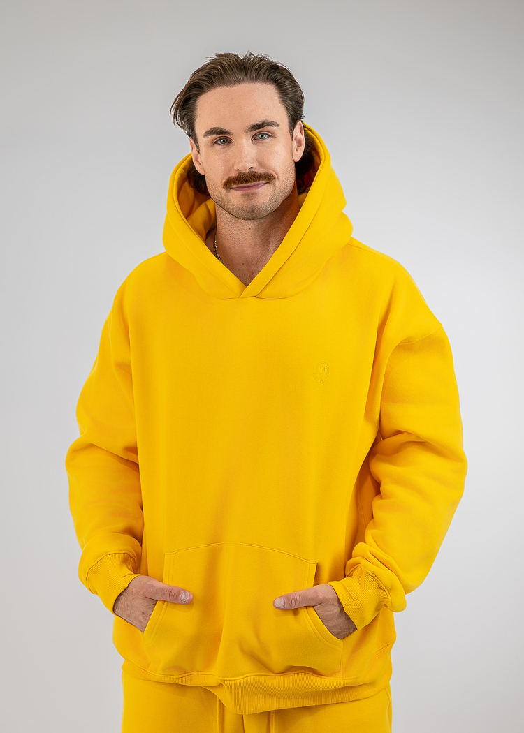 A man wearing a bright yellow pullover hoodie and matching yellow sweatpants, standing against a light gray studio background with his hands in the hoodie's front pockets.