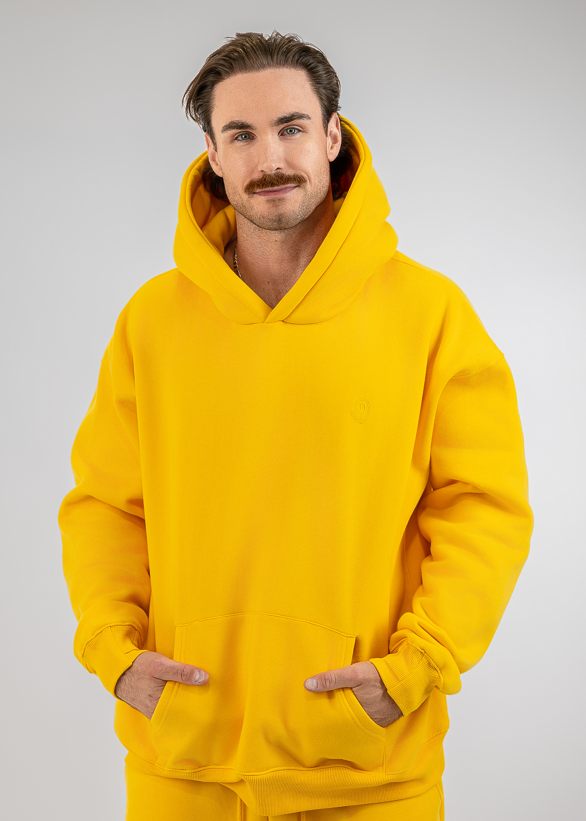 A man wearing a bright yellow pullover hoodie and matching yellow sweatpants, standing against a light gray studio background with his hands in the hoodie's front pockets.