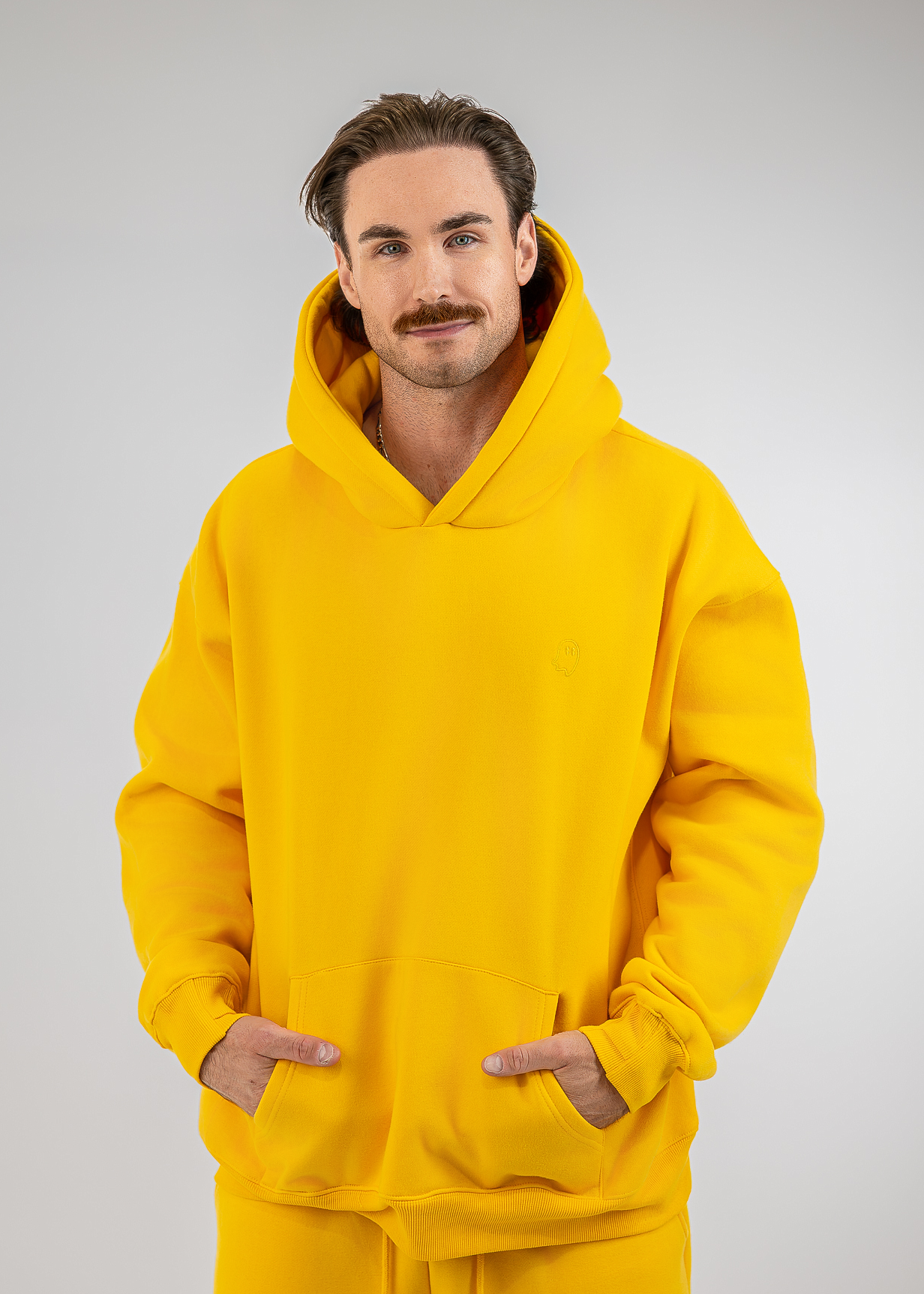 A man wearing a bright yellow pullover hoodie and matching yellow sweatpants, standing against a light gray studio background with his hands in the hoodie's front pockets.