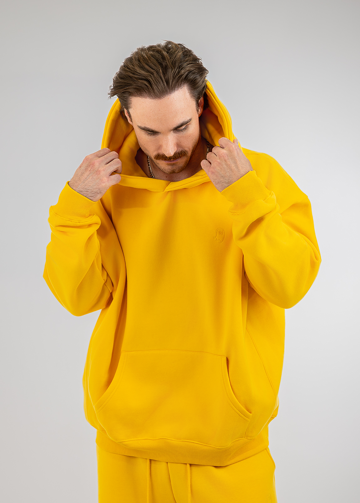 A man wearing a bright yellow matching hoodie and sweatpants set, holding the hood up with both hands against a light gray studio background.