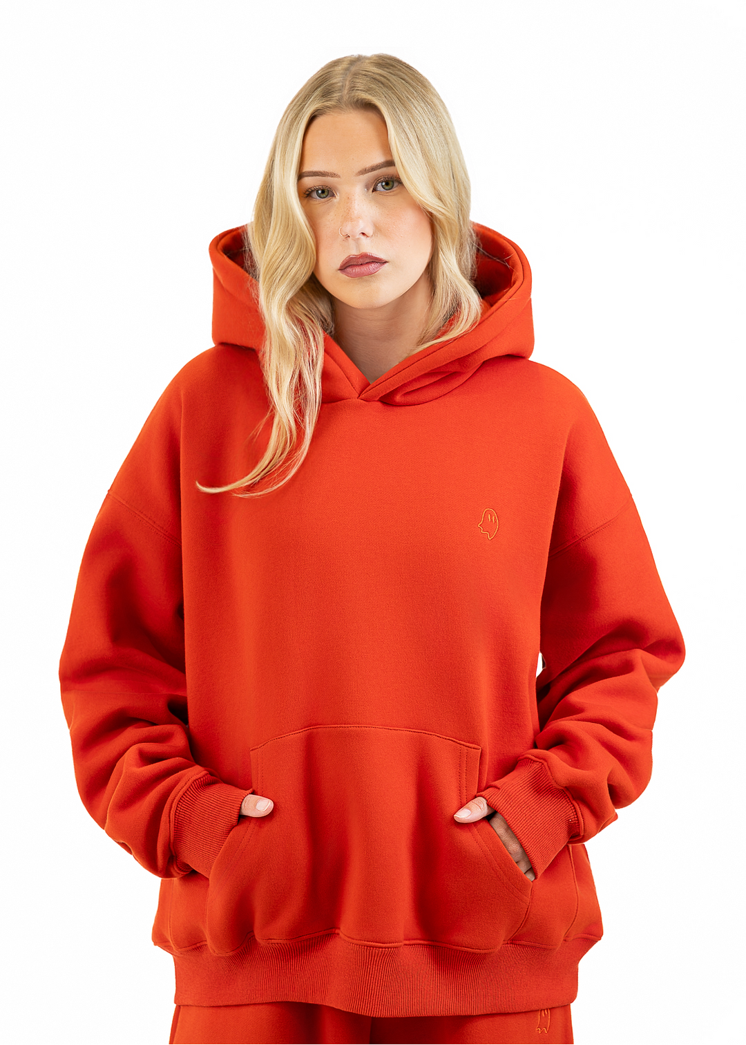 A blonde woman wearing an oversized bright orange-red hoodie with a small tonal embroidered logo on the left chest, paired with matching red sweatpants, against a plain white background.