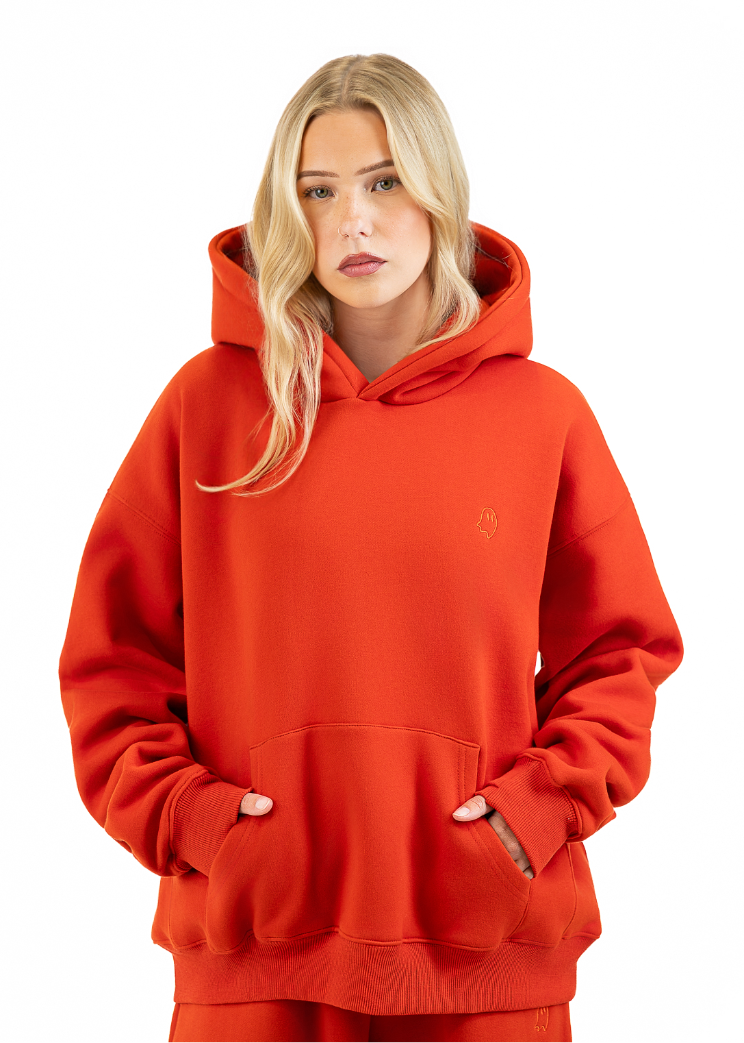 A blonde woman wearing an oversized bright orange-red hoodie with a small tonal embroidered logo on the left chest, paired with matching red sweatpants, against a plain white background.