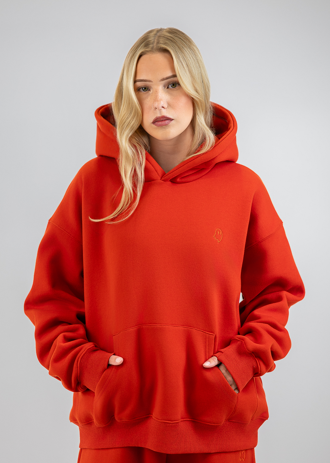 A blonde woman wearing an oversized bright red hoodie and matching red sweatpants, standing against a neutral gray studio background.