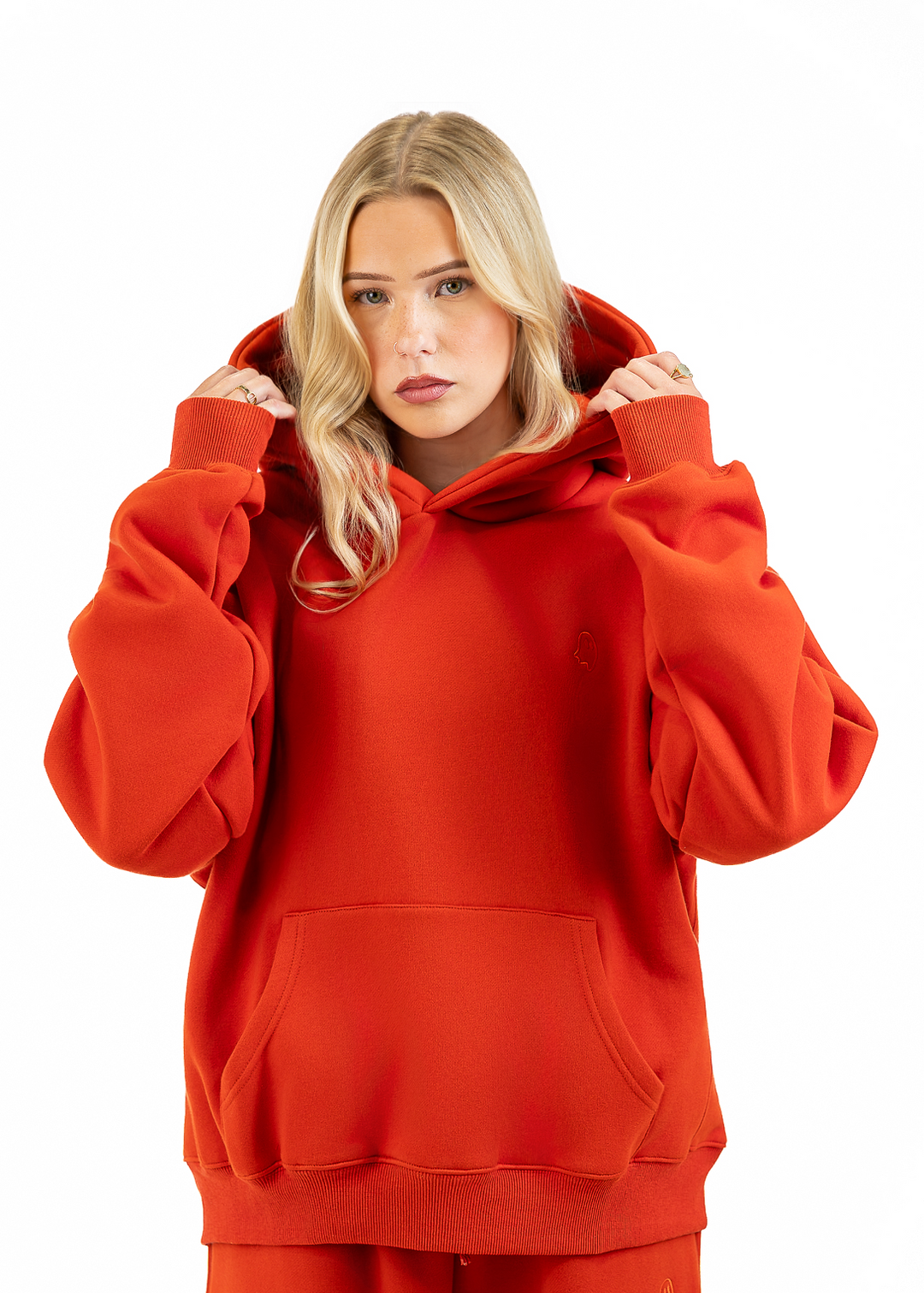 A blonde woman wearing a bright red oversized hoodie and matching red sweatpants, holding the hood up against a plain white studio background.