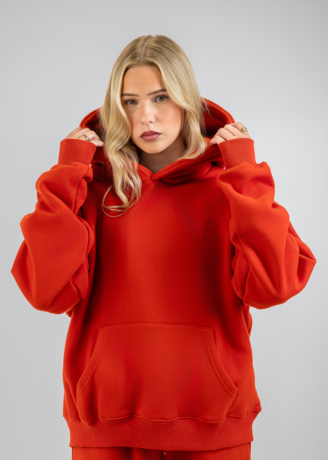 A blonde woman wearing an oversized bright red hoodie and matching red sweatpants, posing against a neutral gray studio background.