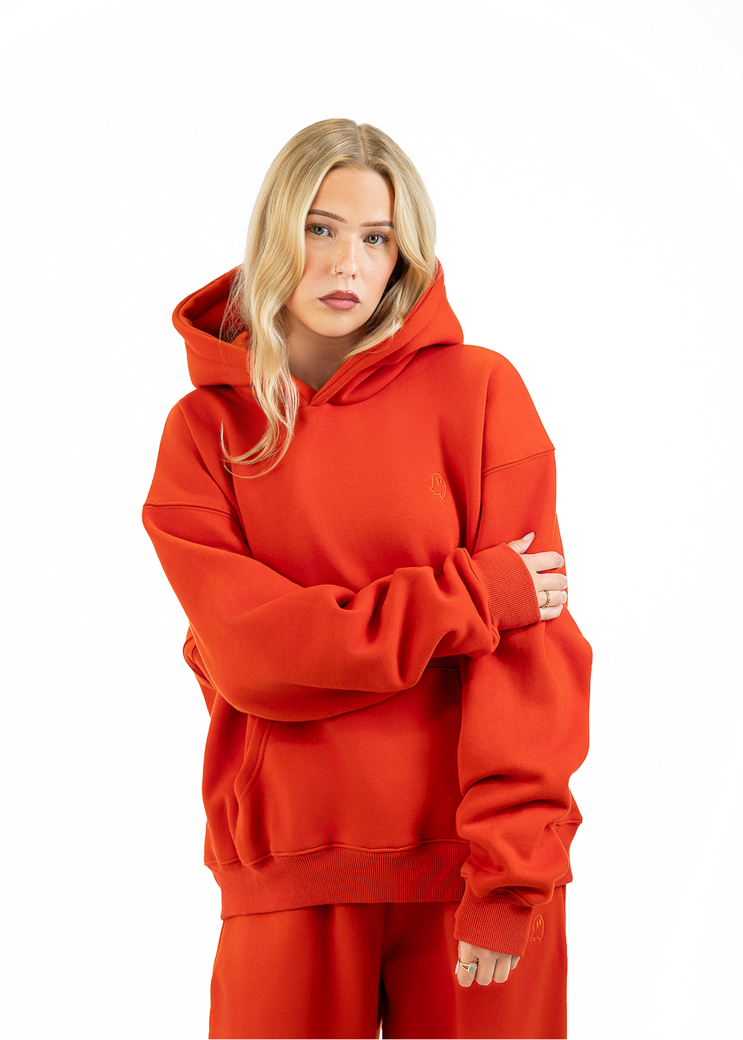 A blonde woman wearing an oversized bright red hoodie and matching red sweatpants, standing against a plain white studio background with her arms crossed.