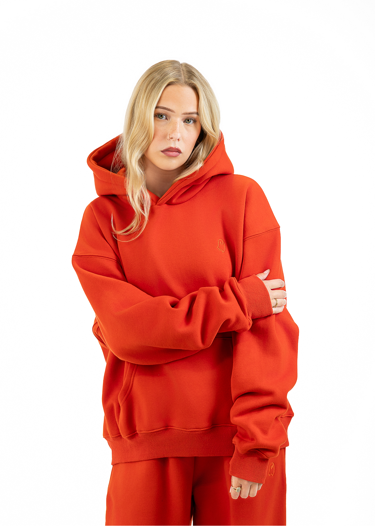 A blonde woman wearing an oversized bright red hoodie and matching red sweatpants, standing against a plain white studio background with her arms crossed.