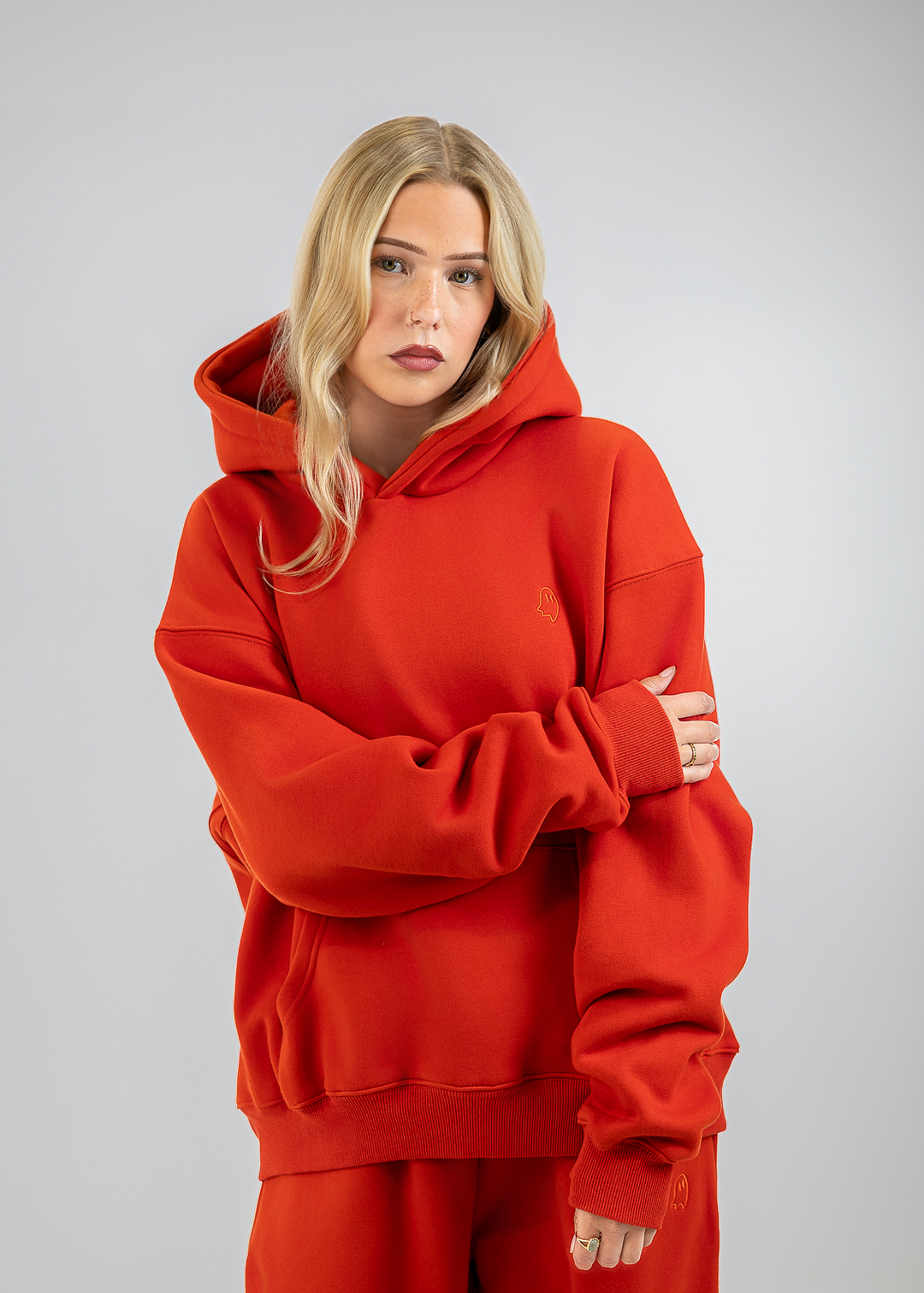A blonde woman wearing a matching red oversized hoodie and sweatpants set, standing against a neutral gray studio background.
