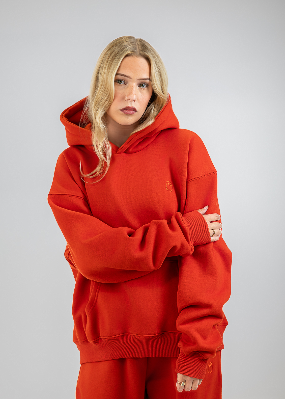 A blonde woman wearing a matching red oversized hoodie and sweatpants set, standing against a neutral gray studio background.