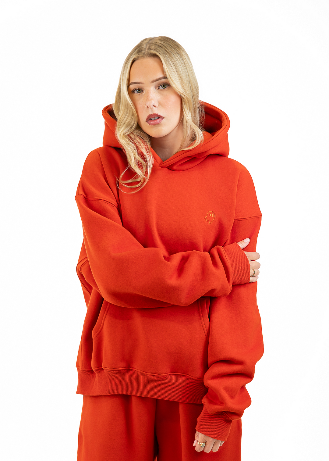 A blonde woman wearing a bright orange-red oversized hoodie and matching sweatpants, standing against a plain white studio background with her arms crossed.