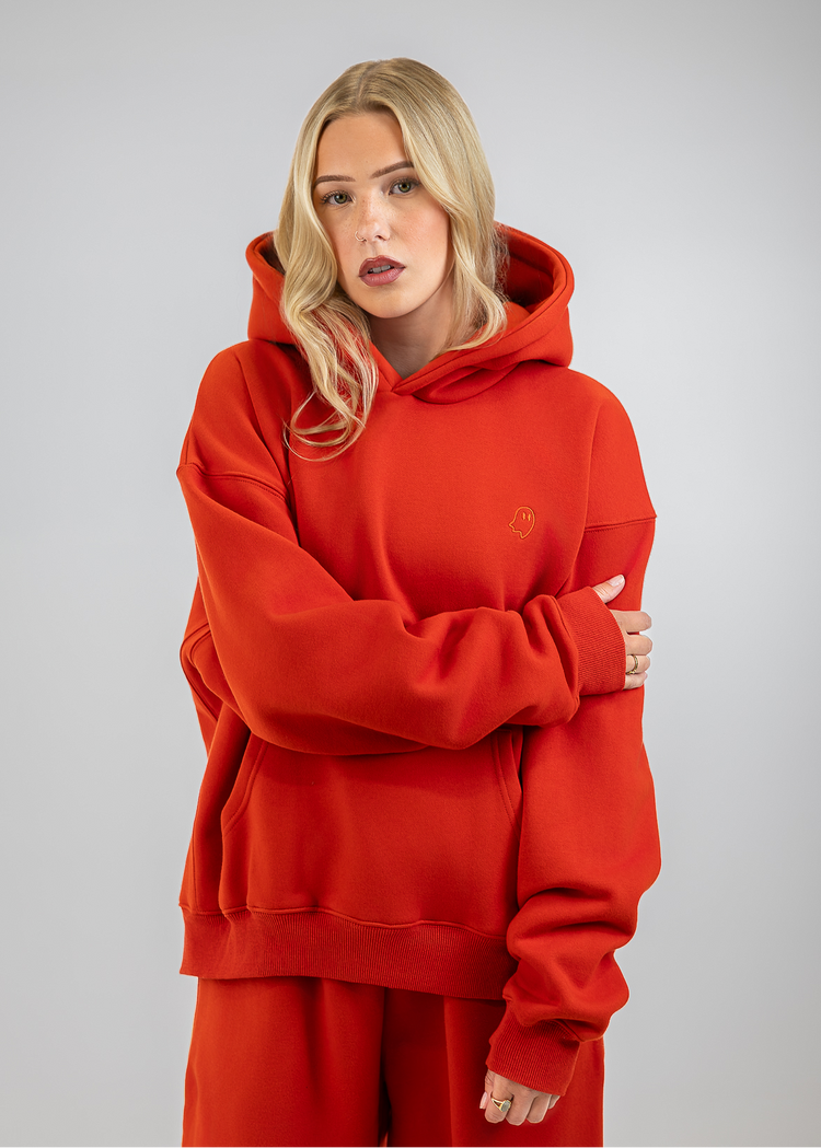 A blonde woman wearing a matching red hoodie and sweatpants set, standing against a neutral gray studio background.