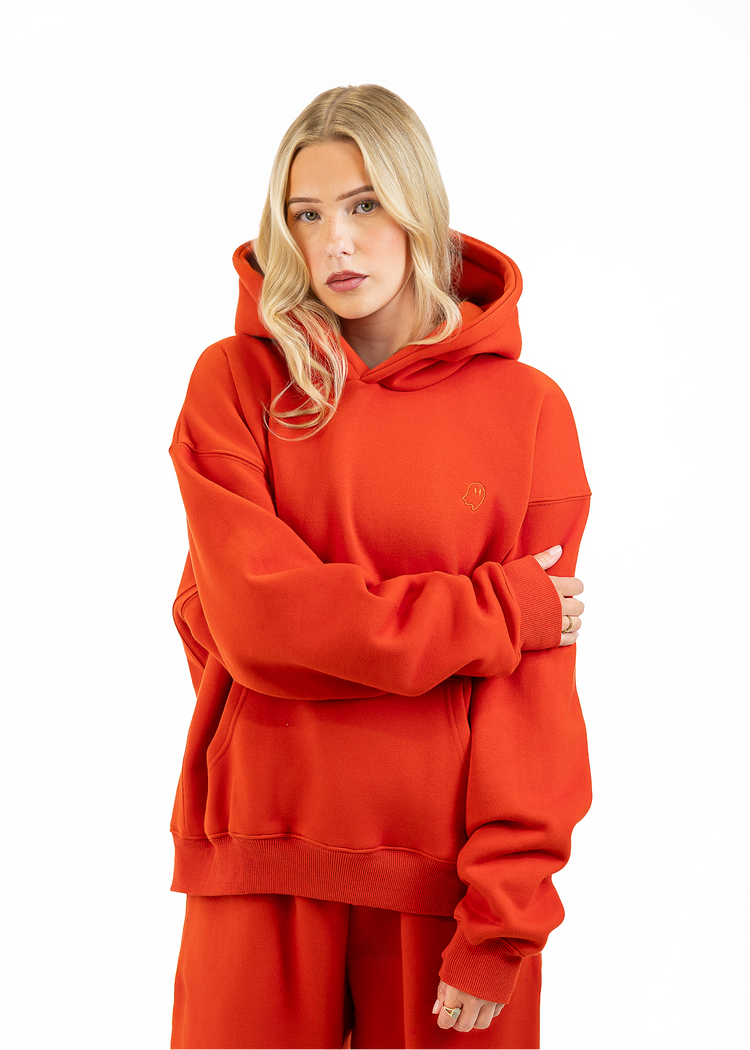 A blonde woman wearing an oversized bright orange-red hooded sweatshirt and matching sweatpants, standing against a plain white studio background with her arms crossed.