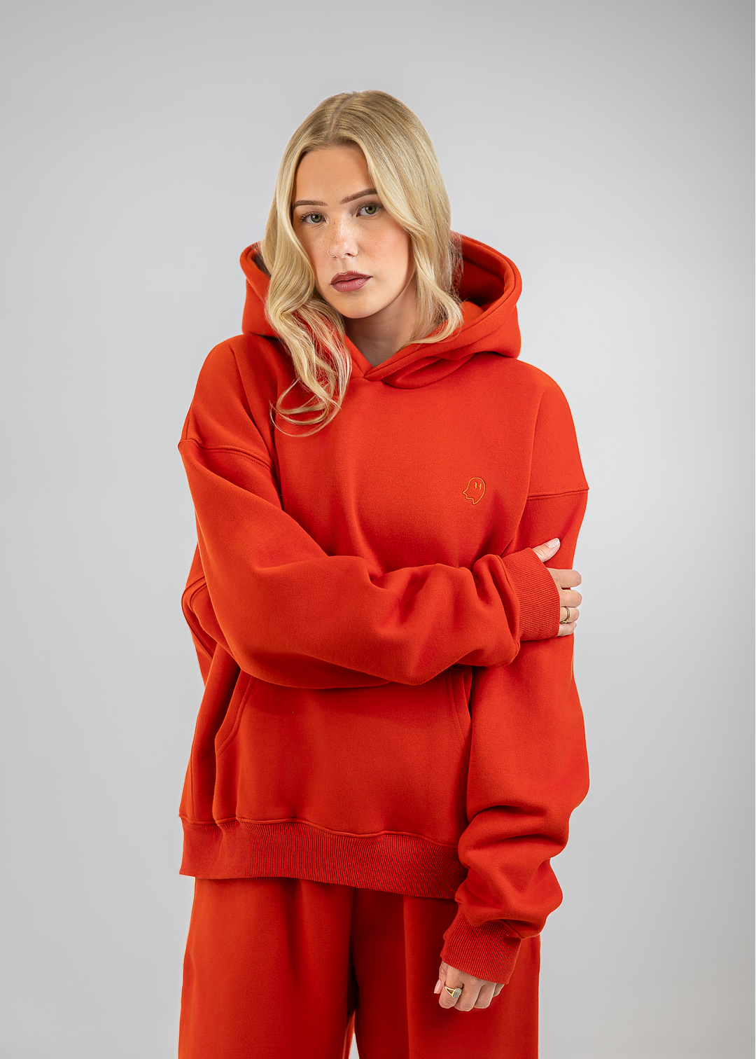 A blonde woman wearing an oversized bright red hoodie and matching red sweatpants, standing against a neutral gray studio background with her arms crossed.