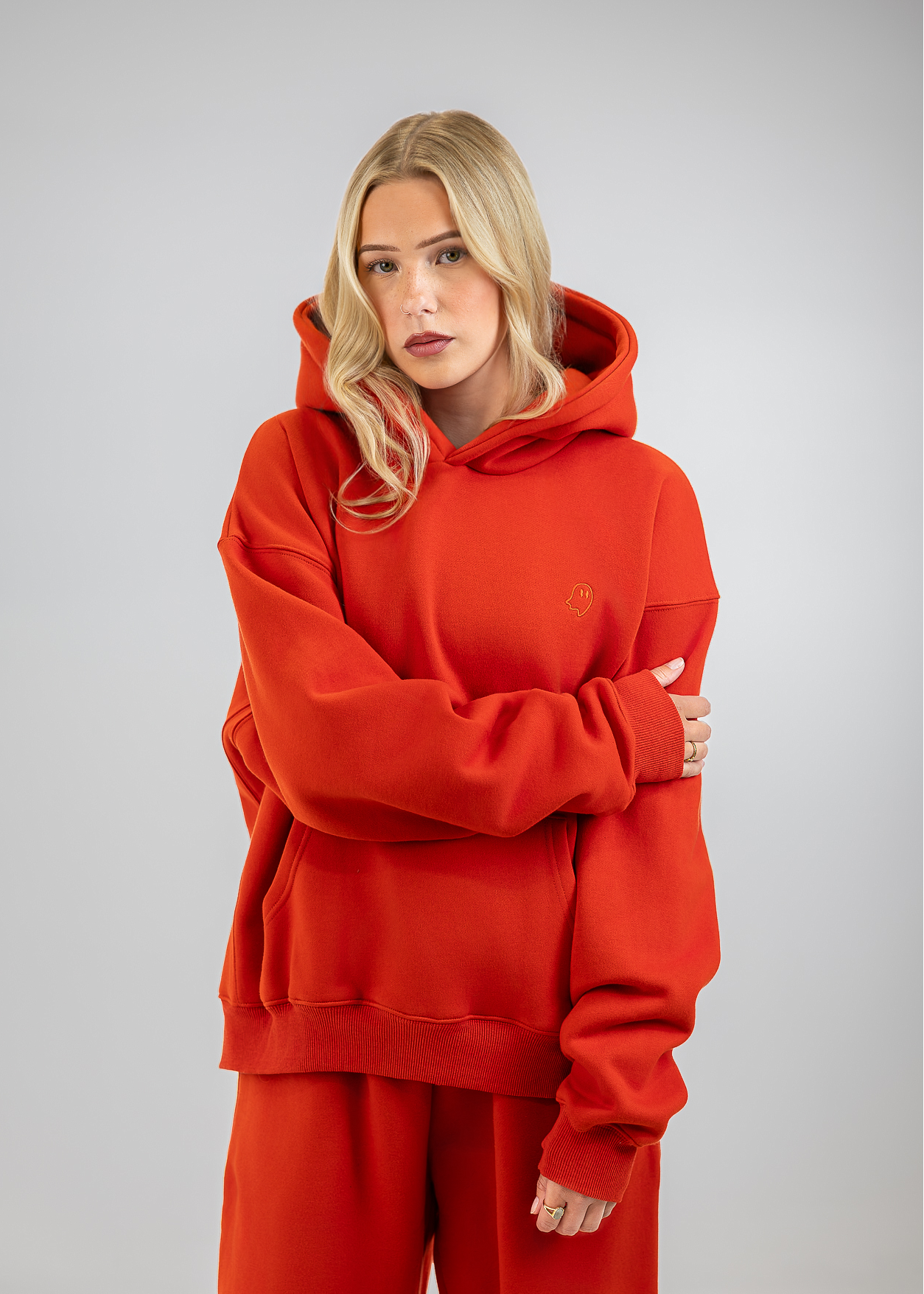 A blonde woman wearing an oversized bright red hoodie and matching red sweatpants, standing against a neutral gray studio background with her arms crossed.