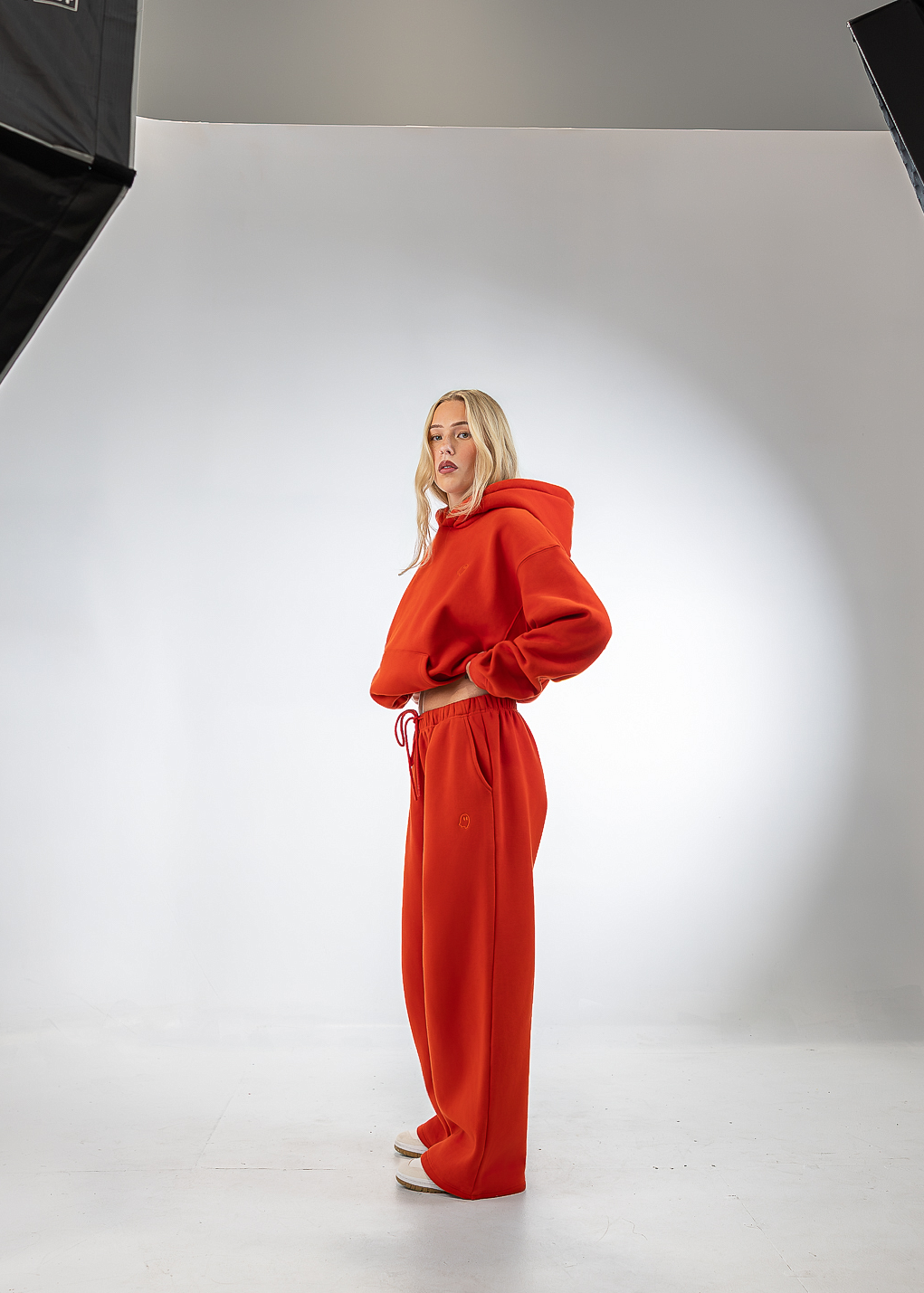 A woman stands in a studio wearing a bright red hooded sweatshirt and matching wide-leg sweatpants. Studio lights are visible on both sides against a white backdrop.