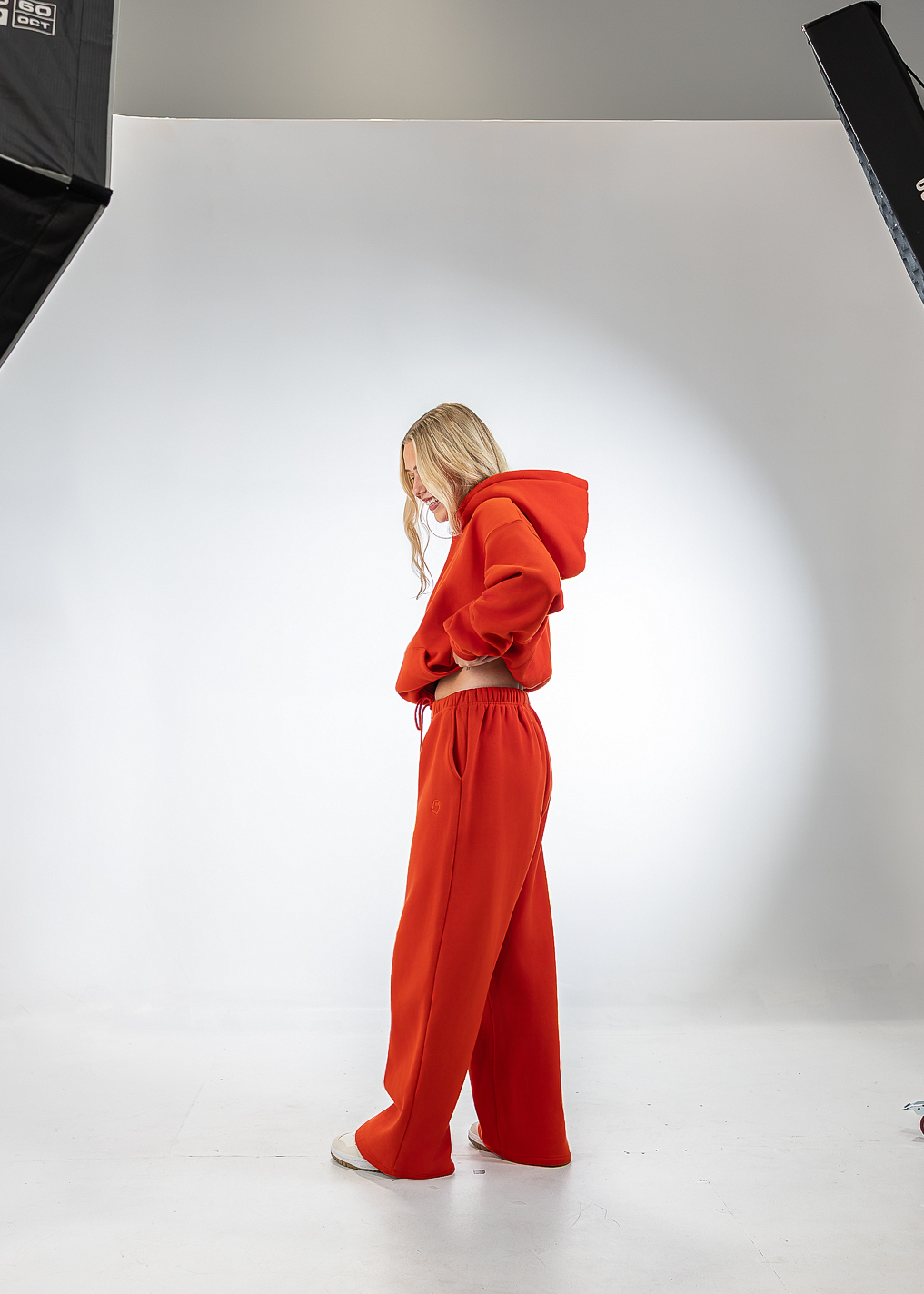 A model wearing a bright red hooded sweatshirt and matching wide-leg sweatpants stands in profile in a white studio setting with softbox lights visible on the left and right.