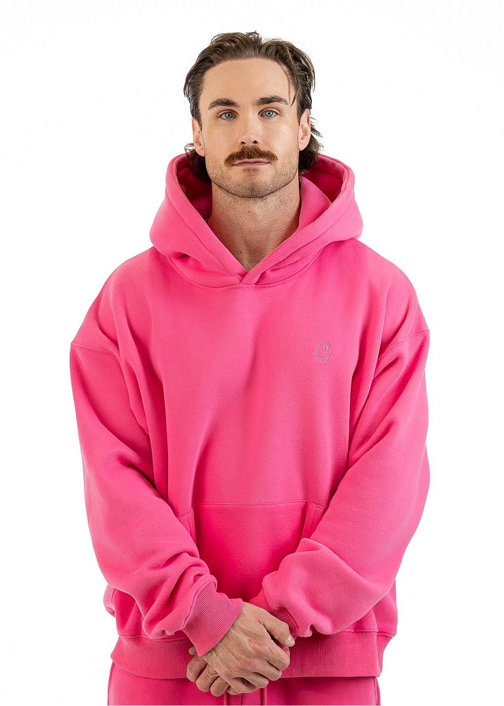 A man wearing a bright pink pullover hoodie and matching pink sweatpants, standing against a plain white studio background.