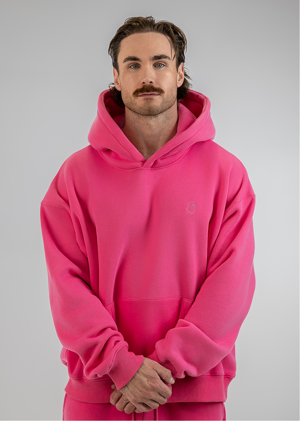 A man wearing a bright pink pullover hoodie and matching pink sweatpants, standing against a neutral gray studio background.