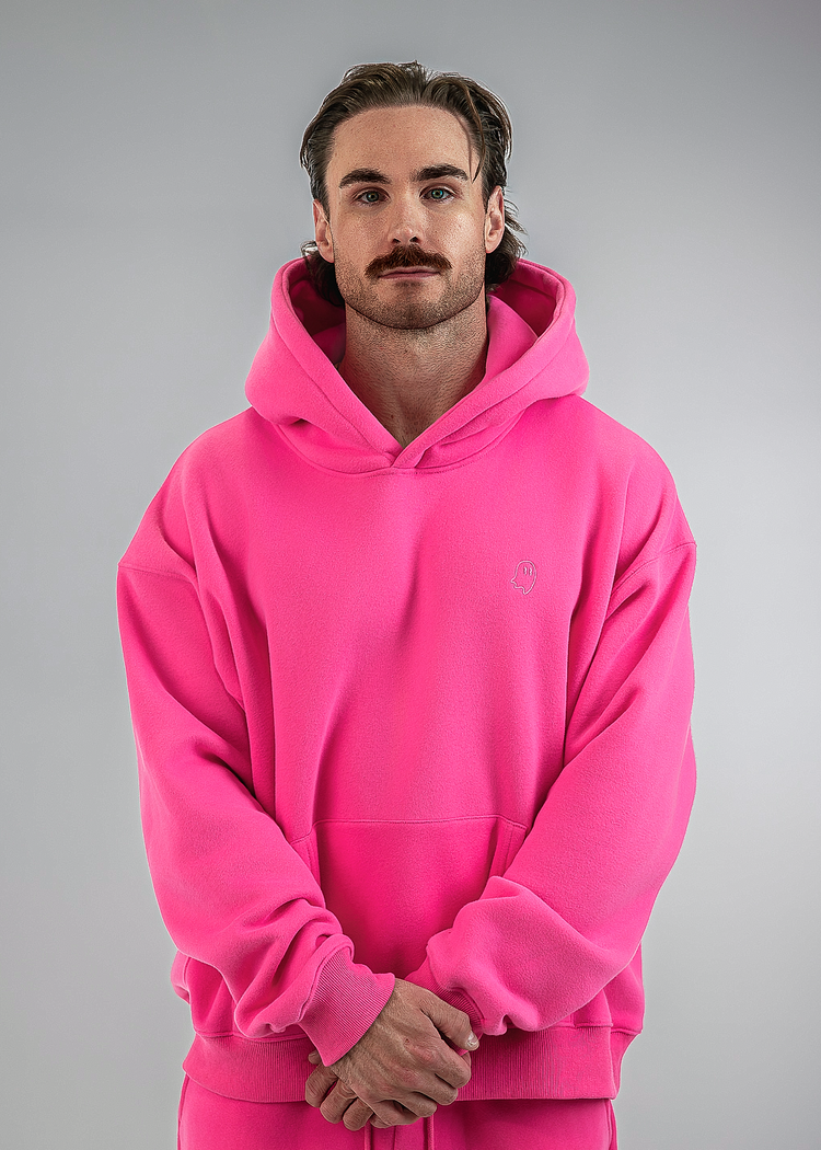 A man wearing a bright pink pullover hoodie and matching pink sweatpants, standing against a neutral gray studio background.