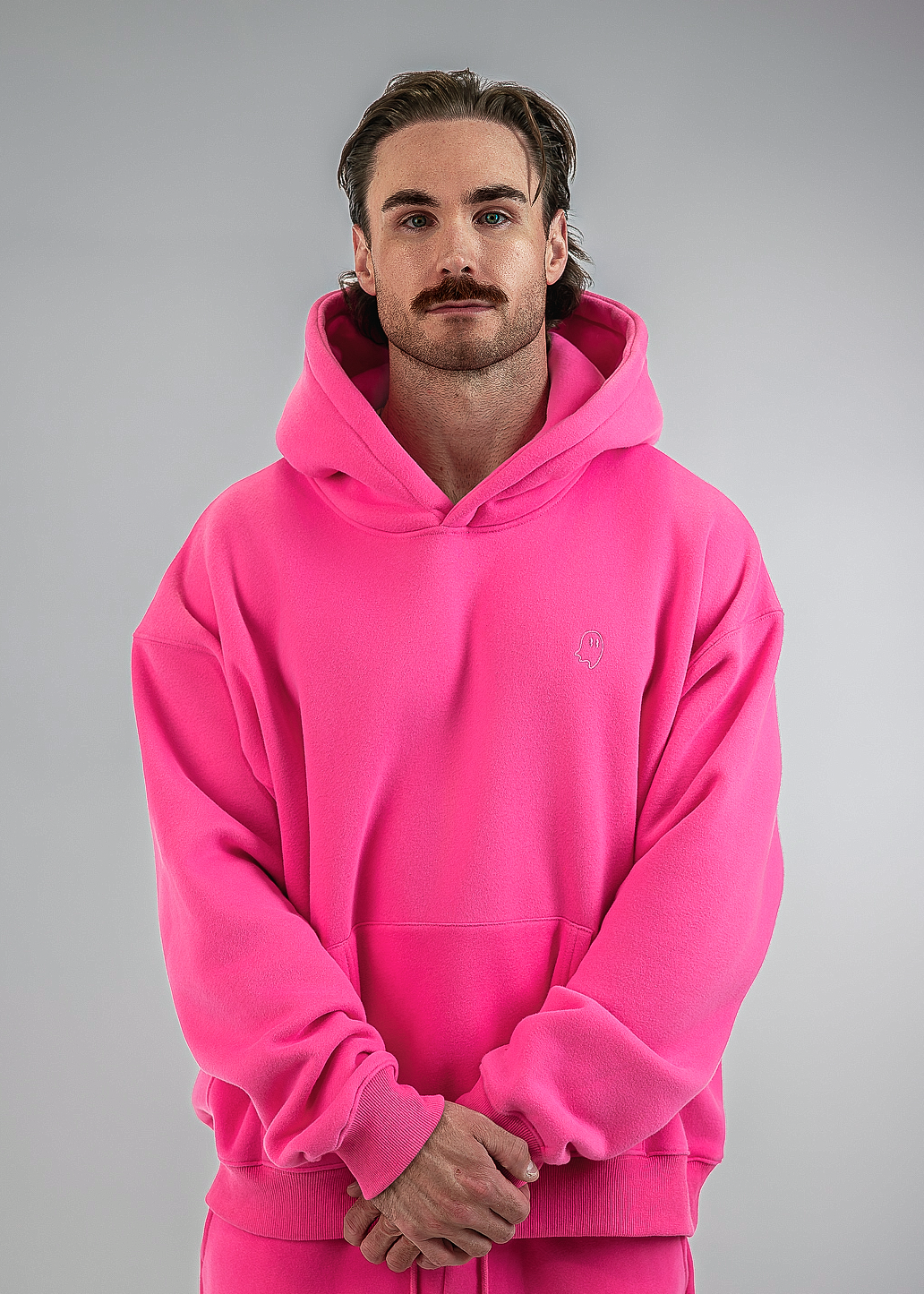 A man wearing a bright pink pullover hoodie and matching pink sweatpants, standing against a neutral gray studio background.