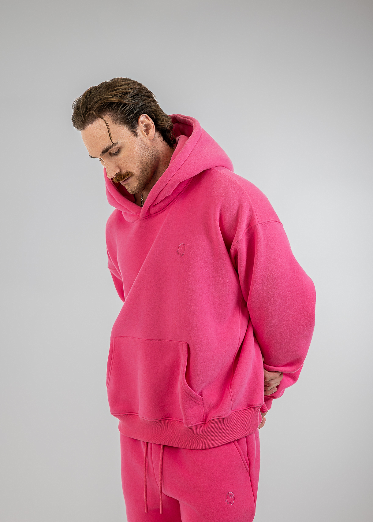 A man wearing a matching hot pink lounge set consisting of an oversized pullover hoodie and jogger pants, photographed against a neutral light gray studio background.