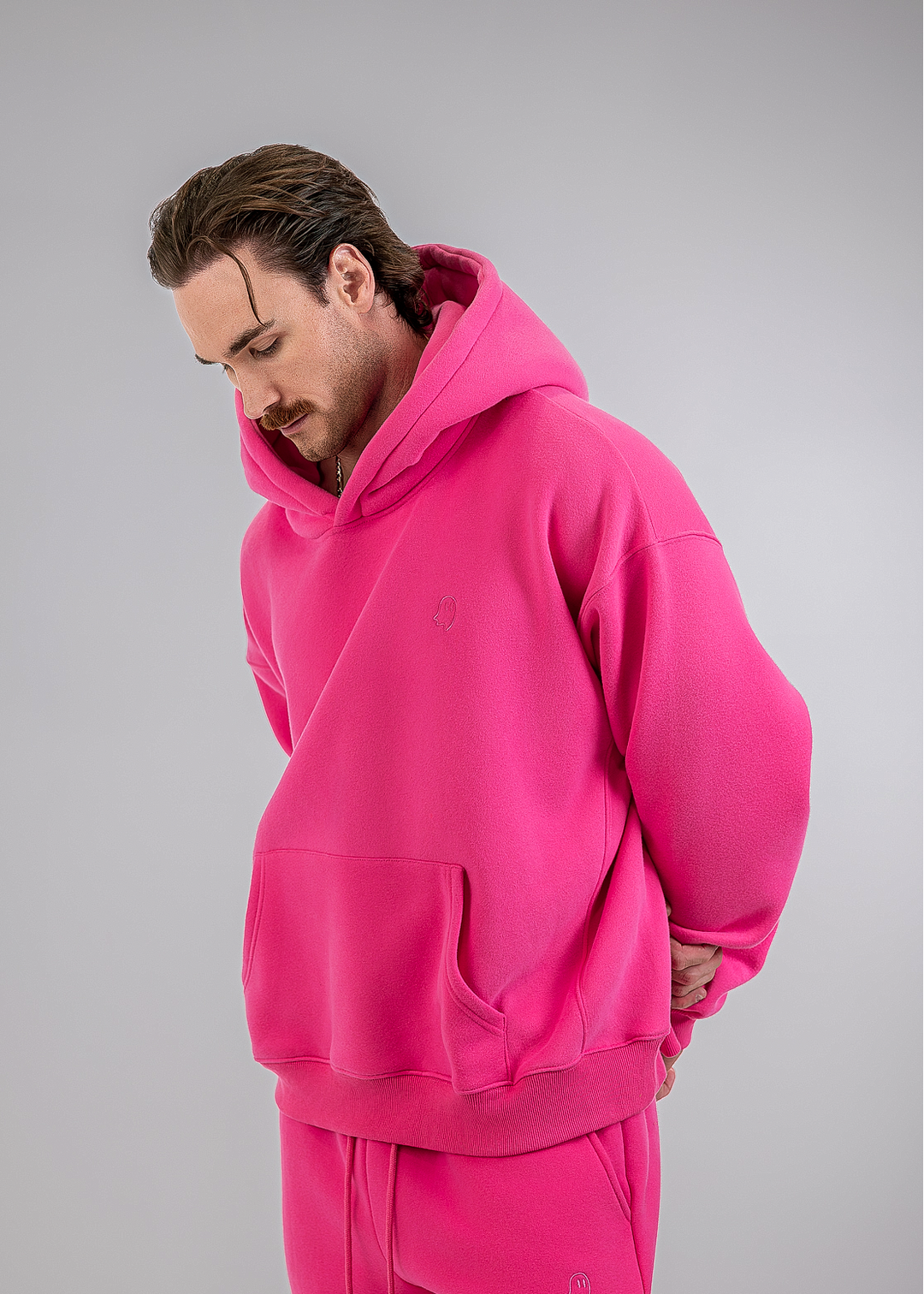 A man wearing a matching hot pink tracksuit, featuring an oversized pullover hoodie and coordinating jogger pants, posed against a neutral gray studio background.