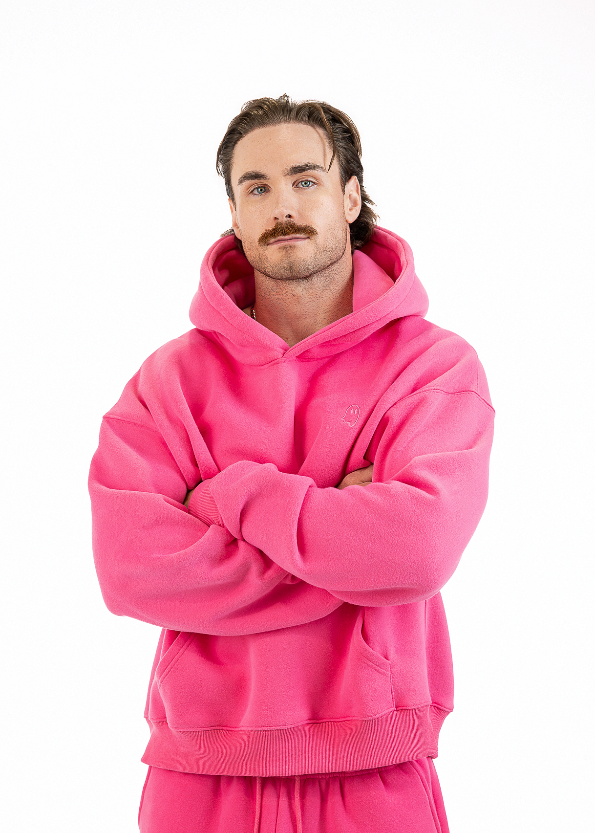 A man with a mustache wearing a bright pink matching hoodie and sweatpants set, standing with arms crossed against a plain white background.