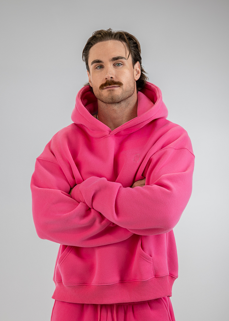 A man with a mustache wearing a bright pink matching hoodie and sweatpants set, standing with his arms crossed against a solid gray studio background.