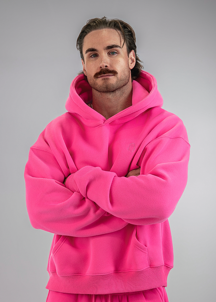 A man wearing a bright pink matching hoodie and sweatpants set, standing with his arms crossed against a solid gray studio background.