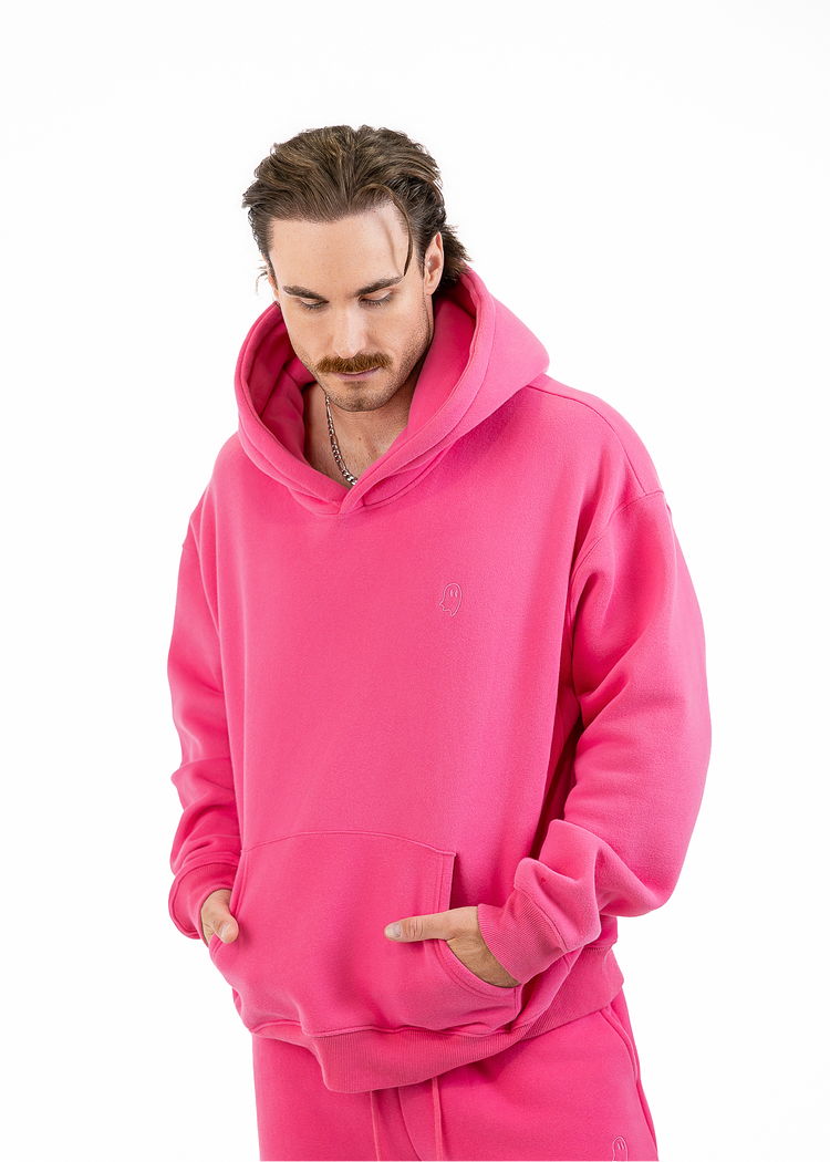 A man wearing a bright pink pullover hoodie and matching pink sweatpants, standing against a plain white studio background.