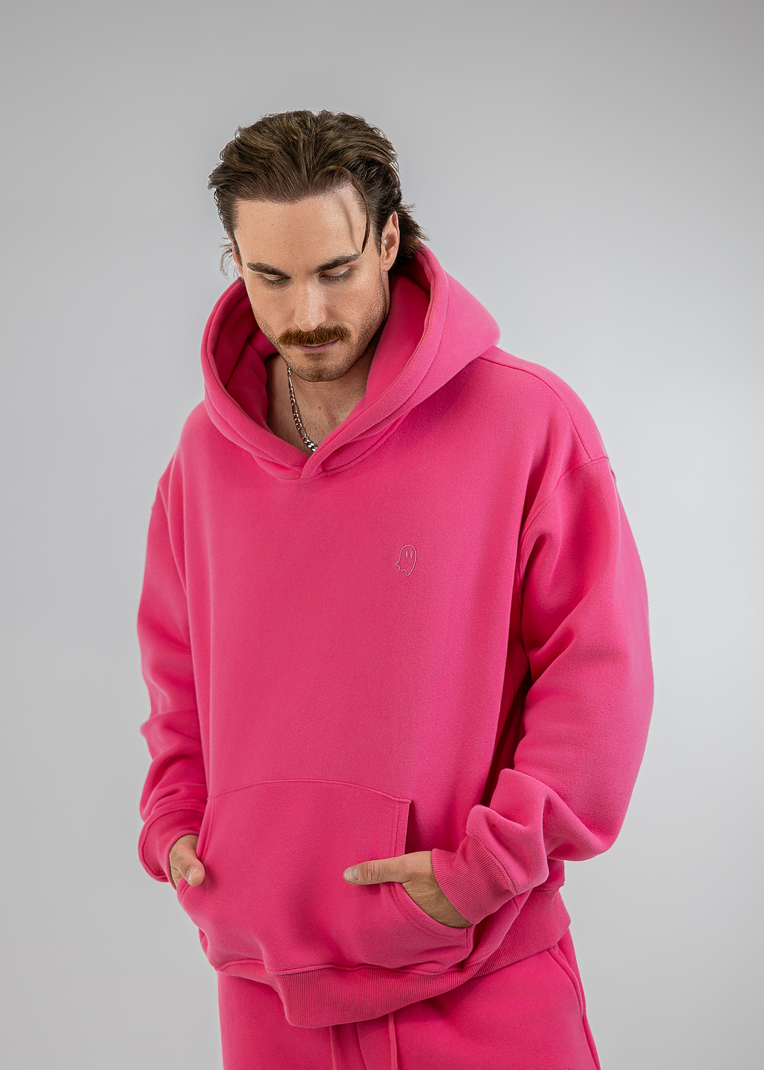 A man wearing a bright pink fleece hoodie and matching sweatpants, standing against a neutral gray studio background.