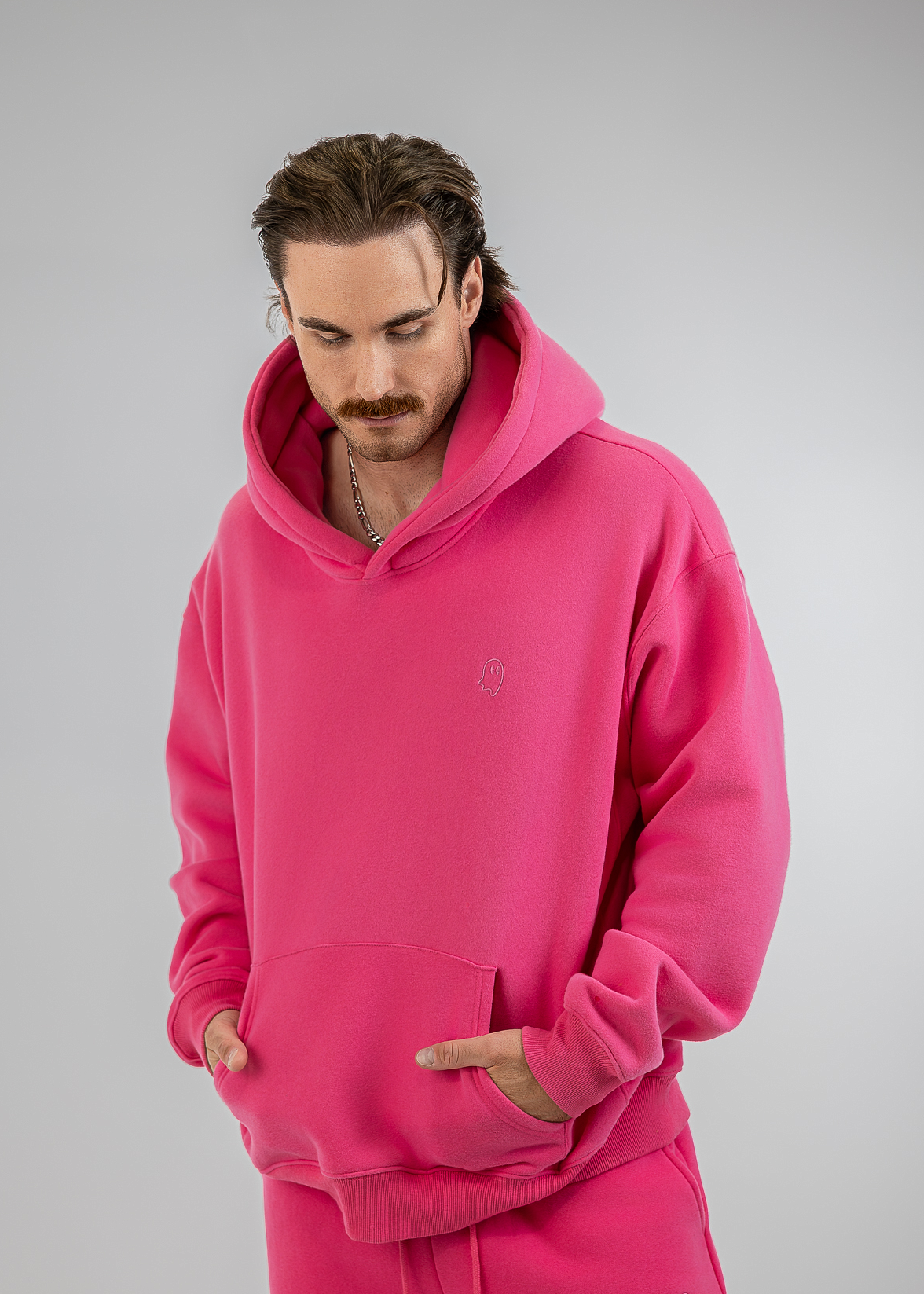 A man wearing a bright pink fleece hoodie and matching sweatpants, standing against a neutral gray studio background.