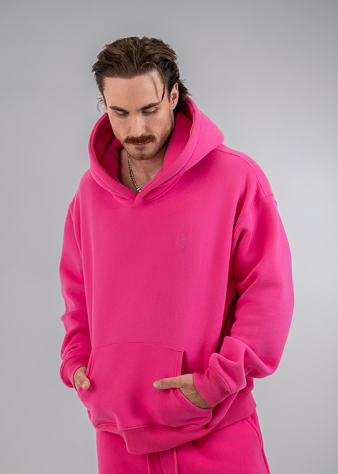 A man wearing a bright pink fleece hoodie and matching sweatpants, standing against a neutral gray studio background.