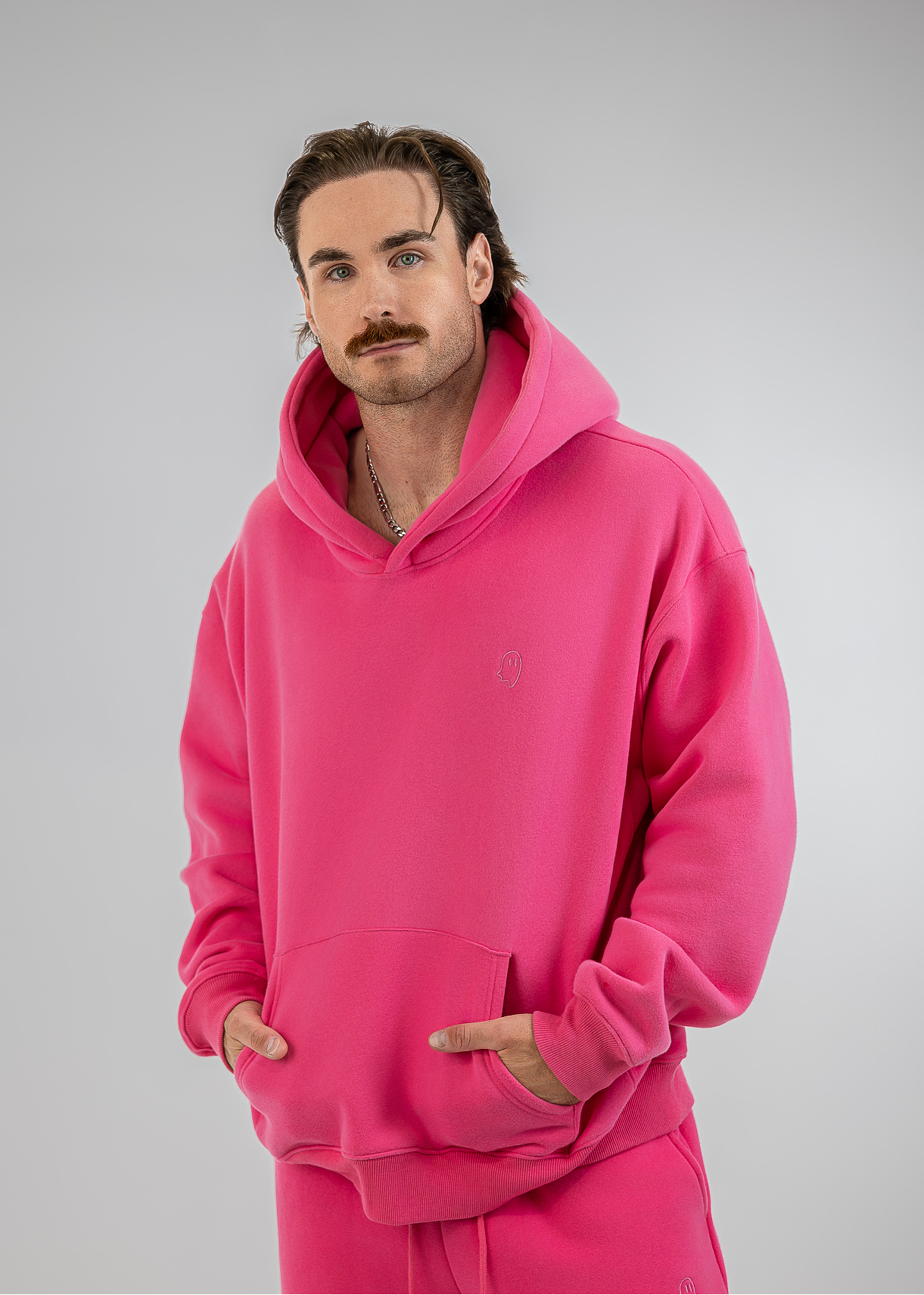 A man wearing a bright pink matching hoodie and sweatpants set, standing against a neutral gray studio background.