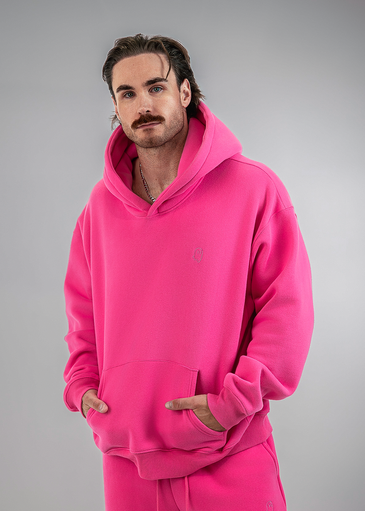 A man wearing a bright pink matching hoodie and sweatpants set, standing against a neutral gray studio background.