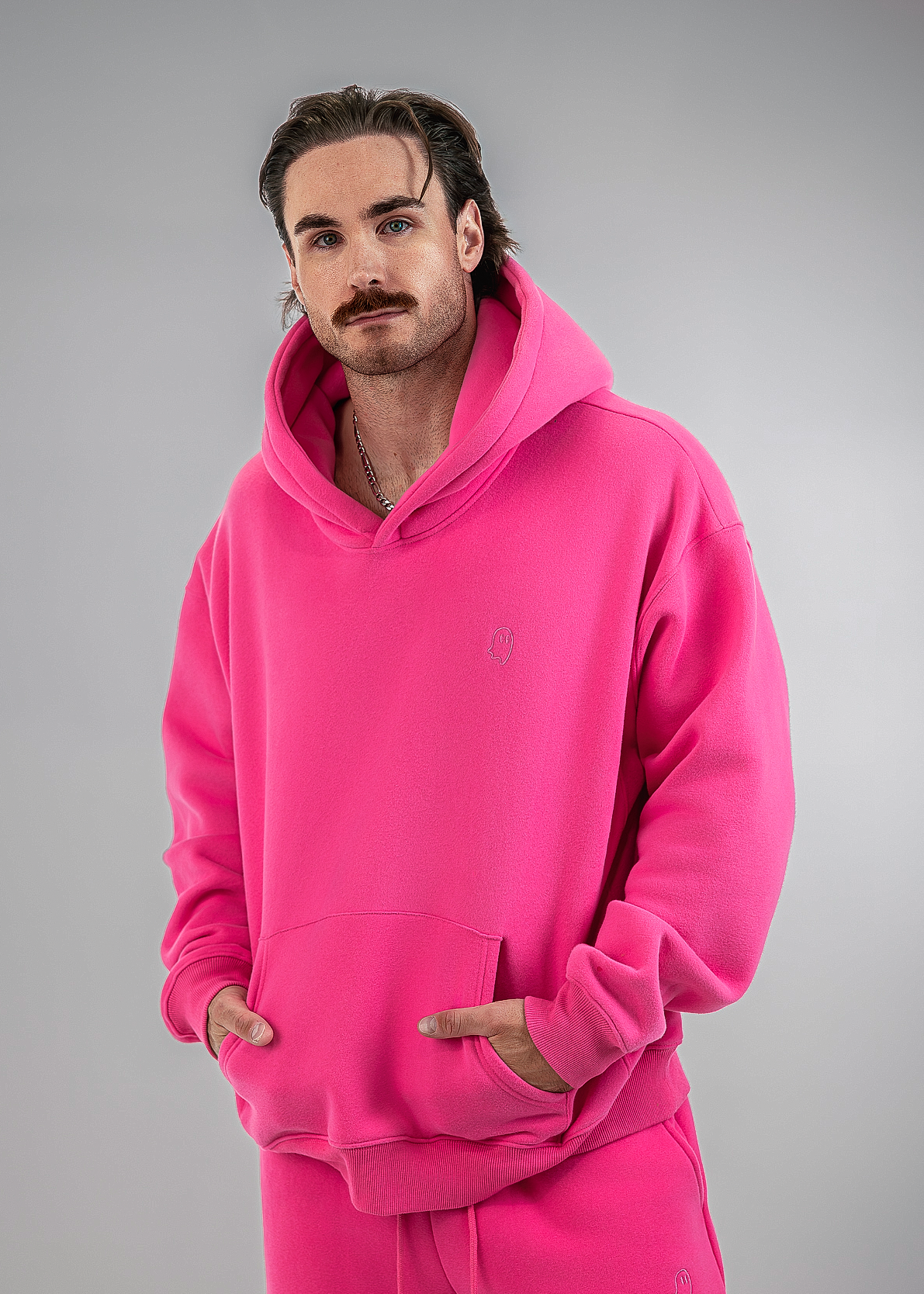 A man wearing a bright pink matching hoodie and sweatpants set, standing against a neutral gray studio background.