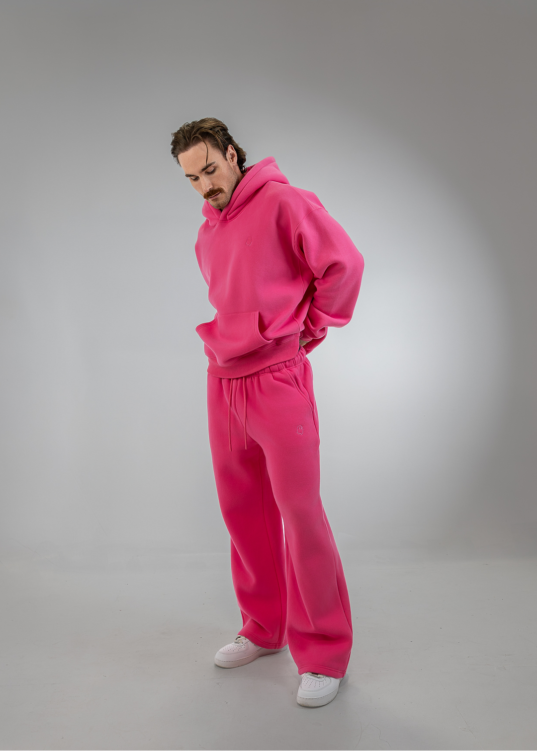A man wearing a bright pink matching hoodie and sweatpants set, styled with white sneakers, standing against a gray studio background.