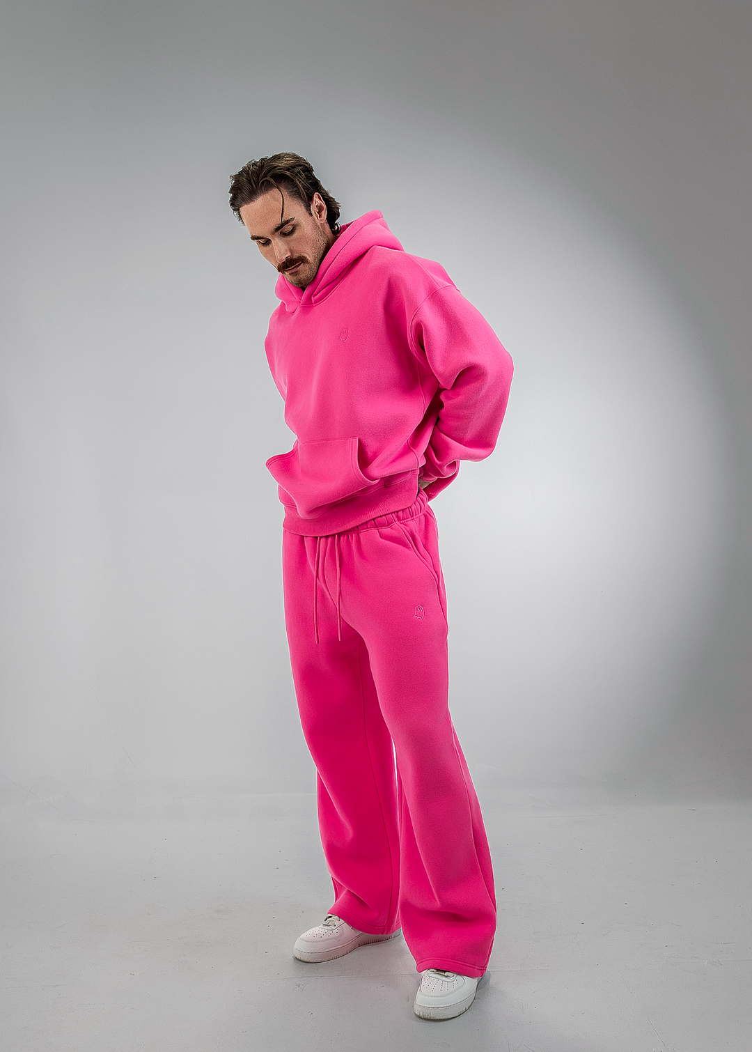 A man wearing a bright pink matching hoodie and sweatpants set, styled with white sneakers, standing in a studio setting with a gray background.