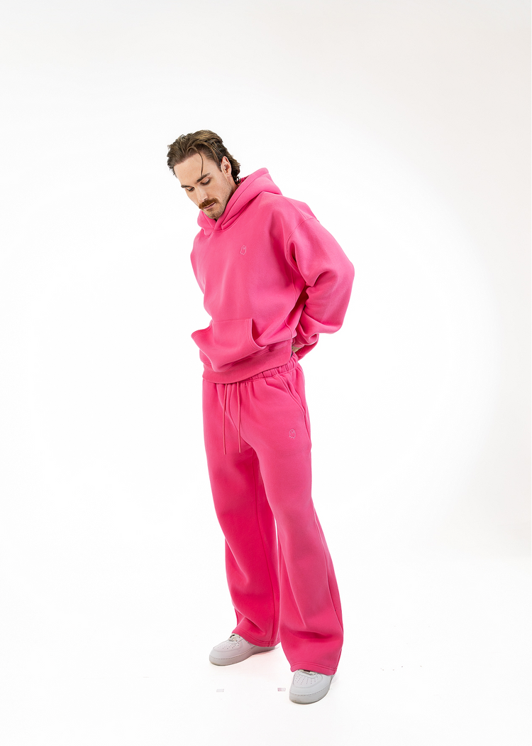 A man wearing a bright pink matching hoodie and sweatpants set, standing against a plain white studio background.