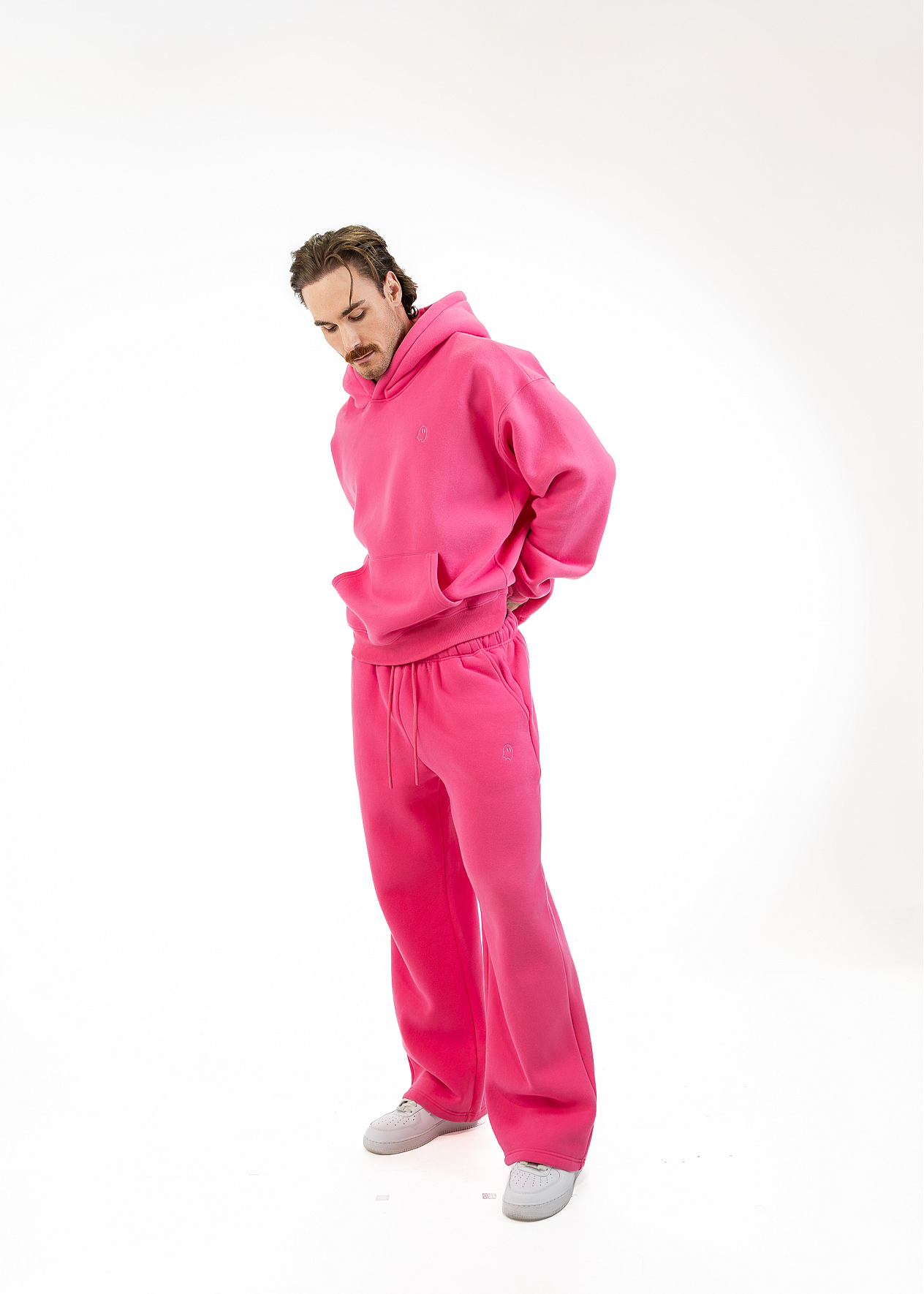A man wearing a bright pink matching hoodie and sweatpants set, standing against a plain white studio background.
