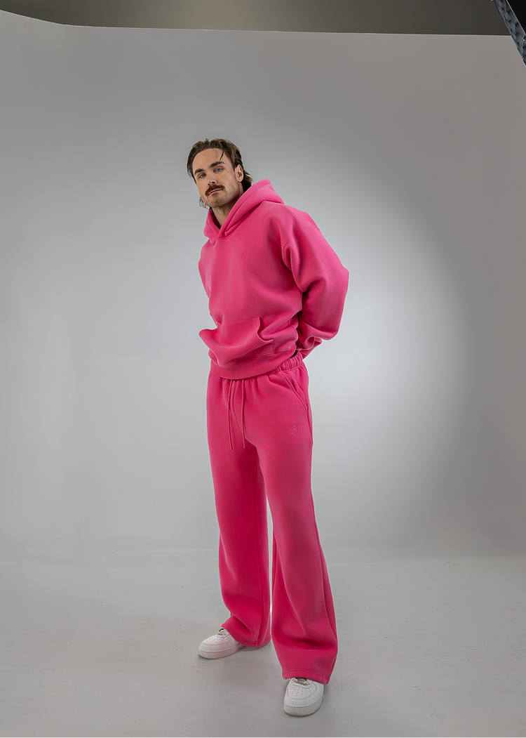 A man modeling a bright pink matching lounge set consisting of a pullover hoodie and wide-leg sweatpants, paired with white sneakers, set against a white studio backdrop.