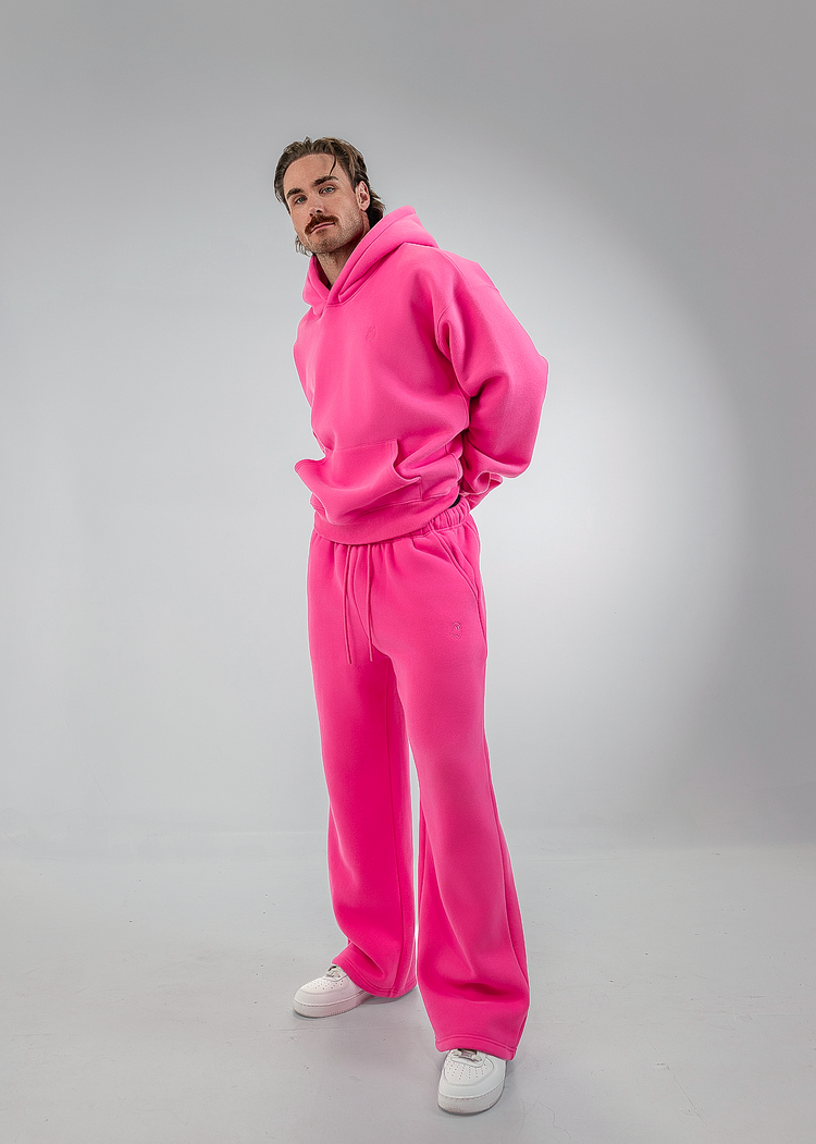 A man modeling a bright pink matching hoodie and sweatpants set with white sneakers, standing in a white studio setting.