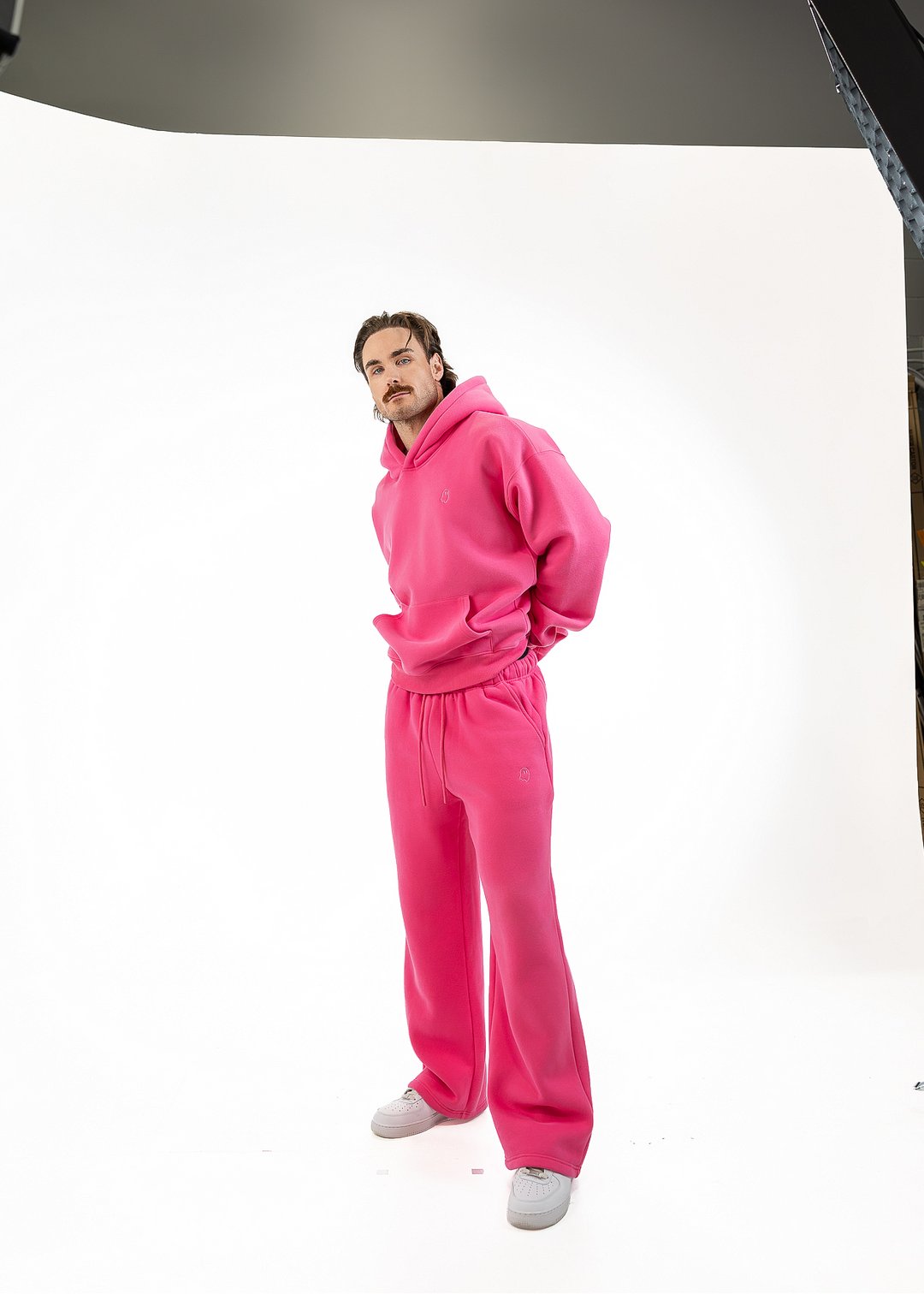 A man modeling a bright pink fleece tracksuit consisting of a pullover hoodie and matching wide-leg sweatpants, standing in a white studio setting.