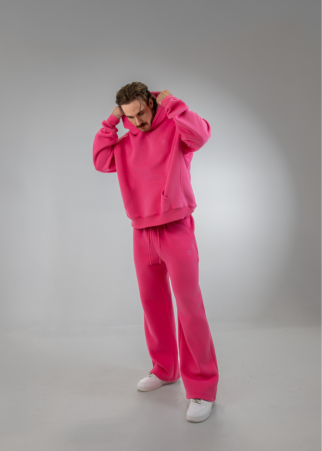 A man modeling a bright pink two-piece tracksuit consisting of a pullover hoodie and matching sweatpants, paired with white sneakers, against a gray studio background.