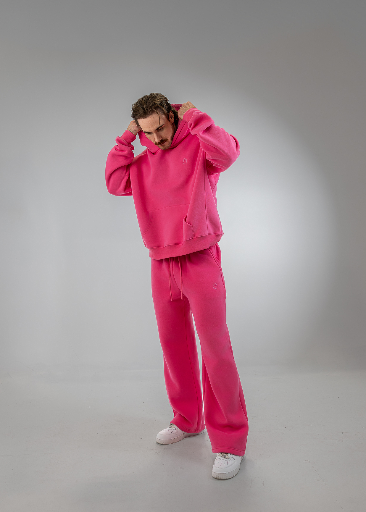 A man modeling a bright pink two-piece tracksuit consisting of a pullover hoodie and matching sweatpants, paired with white sneakers, against a gray studio background.