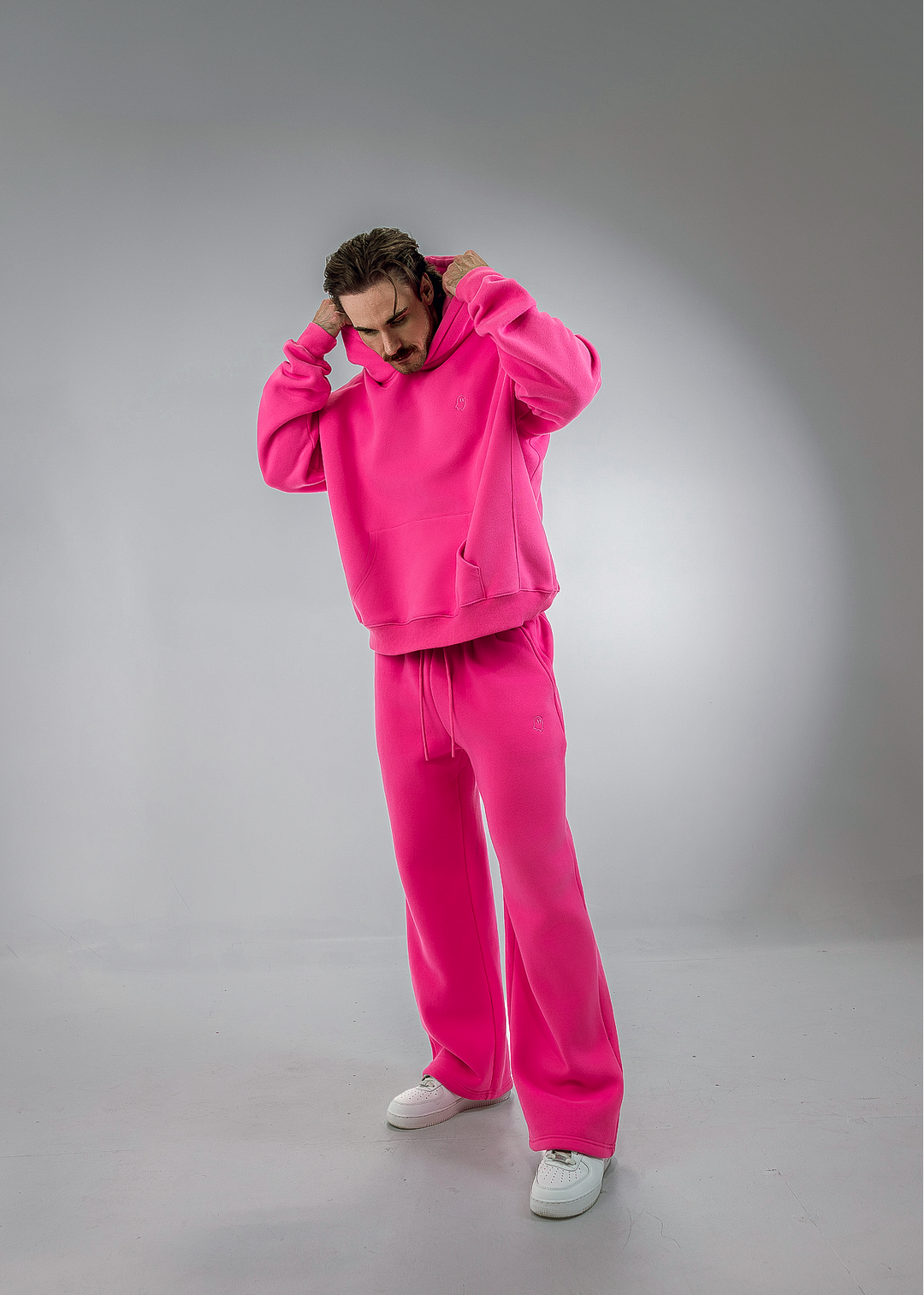 A man modeling a bright pink two-piece tracksuit consisting of a pullover hoodie and matching sweatpants, paired with white sneakers, against a gray studio backdrop.