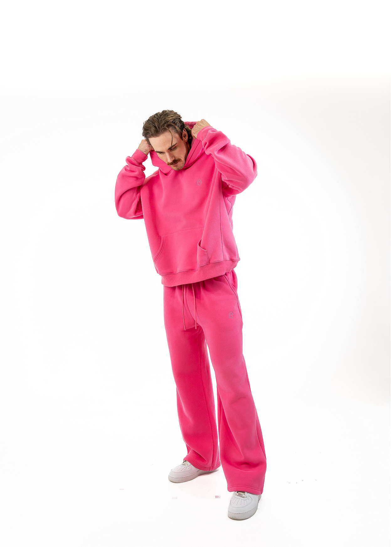 A man wearing a bright pink two-piece tracksuit consisting of a pullover hoodie and matching sweatpants, standing against a solid white background.