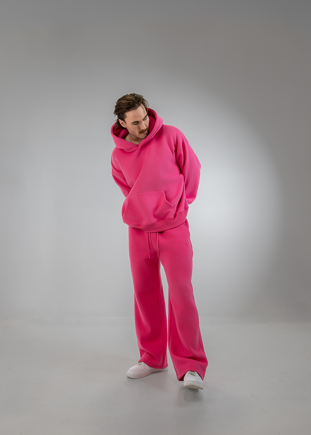 A man wearing a matching bright pink lounge set consisting of a pullover hoodie and wide-leg sweatpants, paired with white sneakers, standing against a gray studio backdrop.
